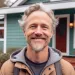 An older man wearing a sweatshirt in his 50's standing outside his home smiling